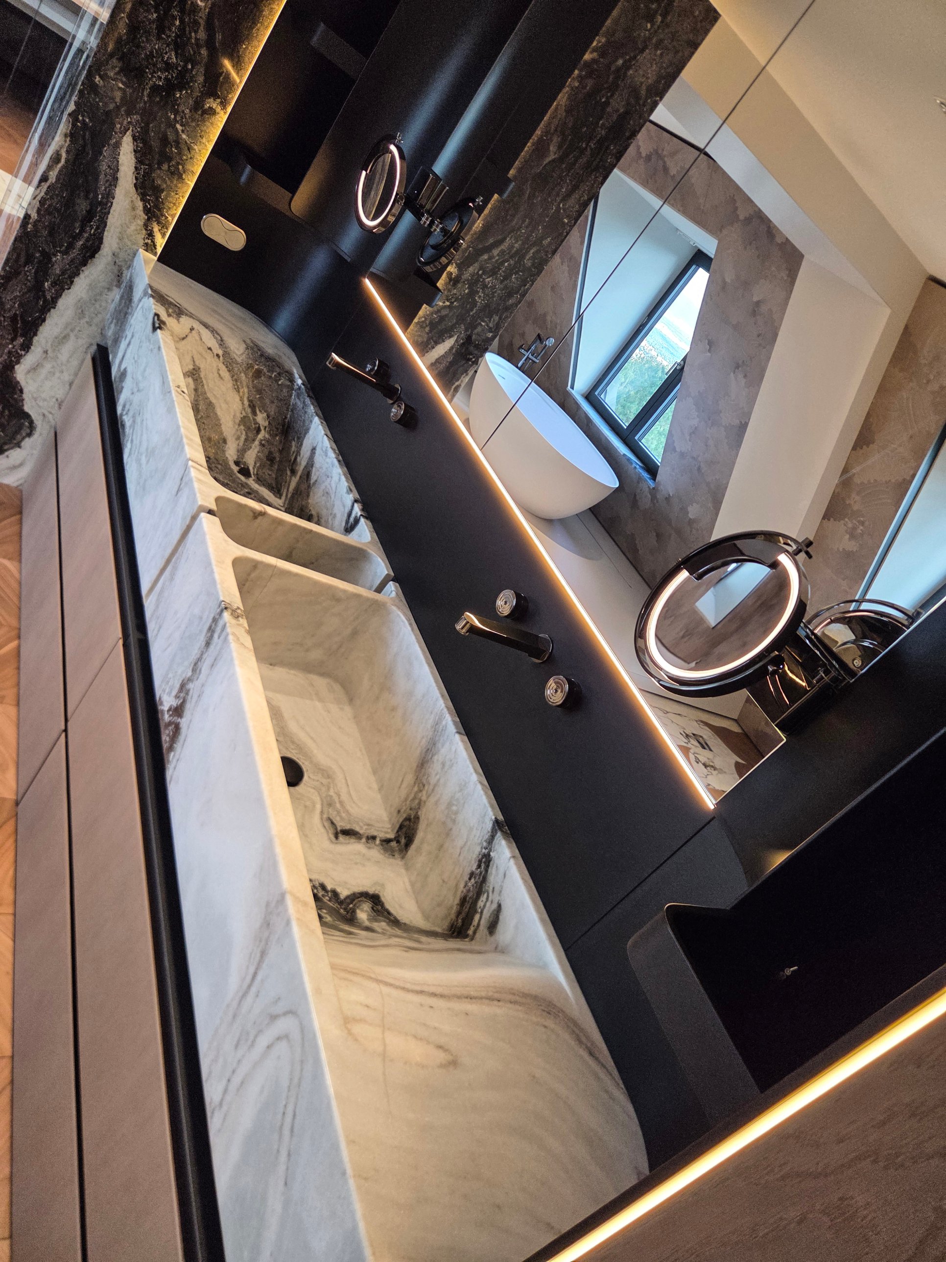 A double stone basin with dark veining, black fittings and a lit mirror reflecting the bath