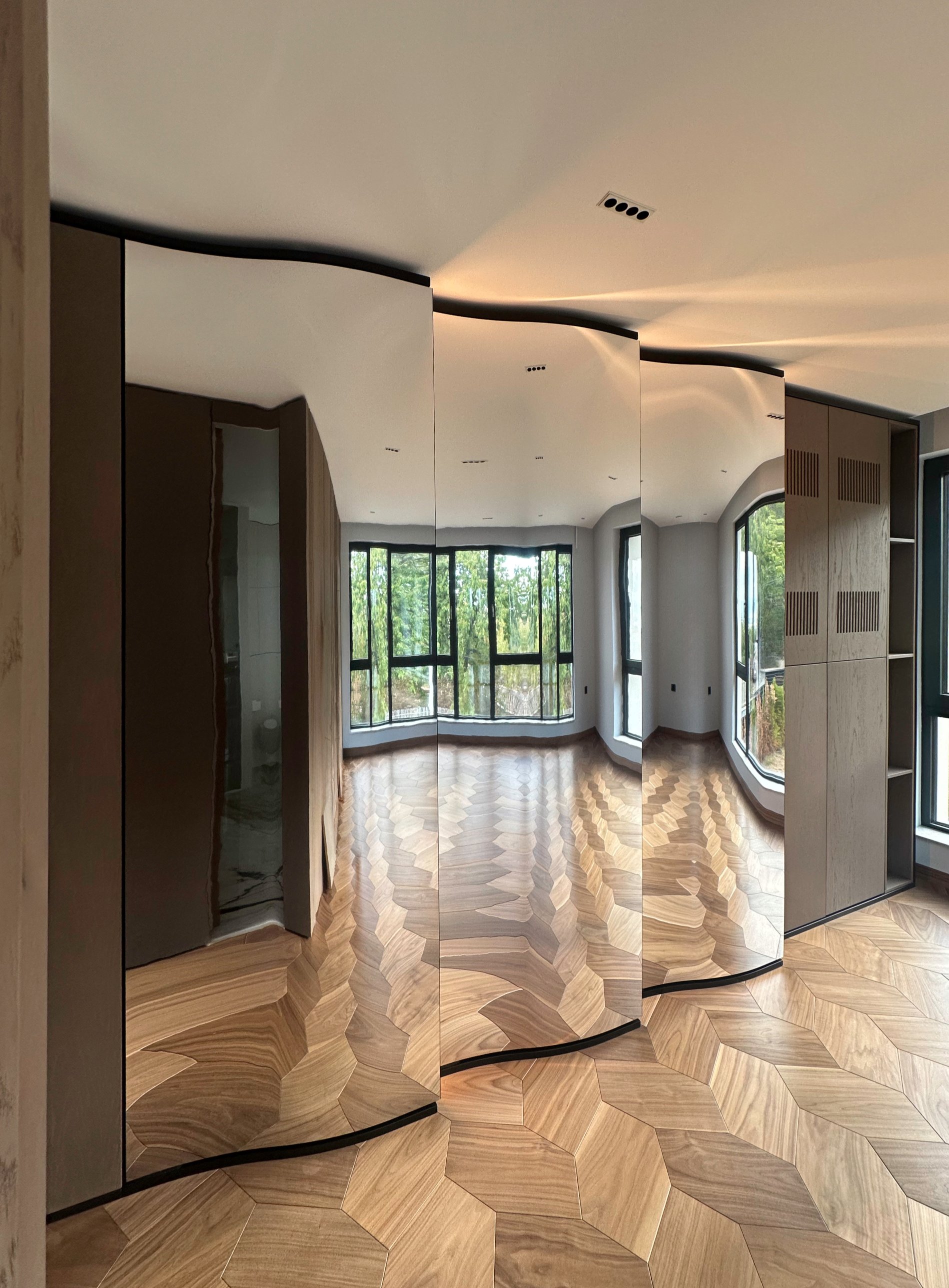 A mirrored wardrobe bending into an undulating wall that reflects the windows and timber floor