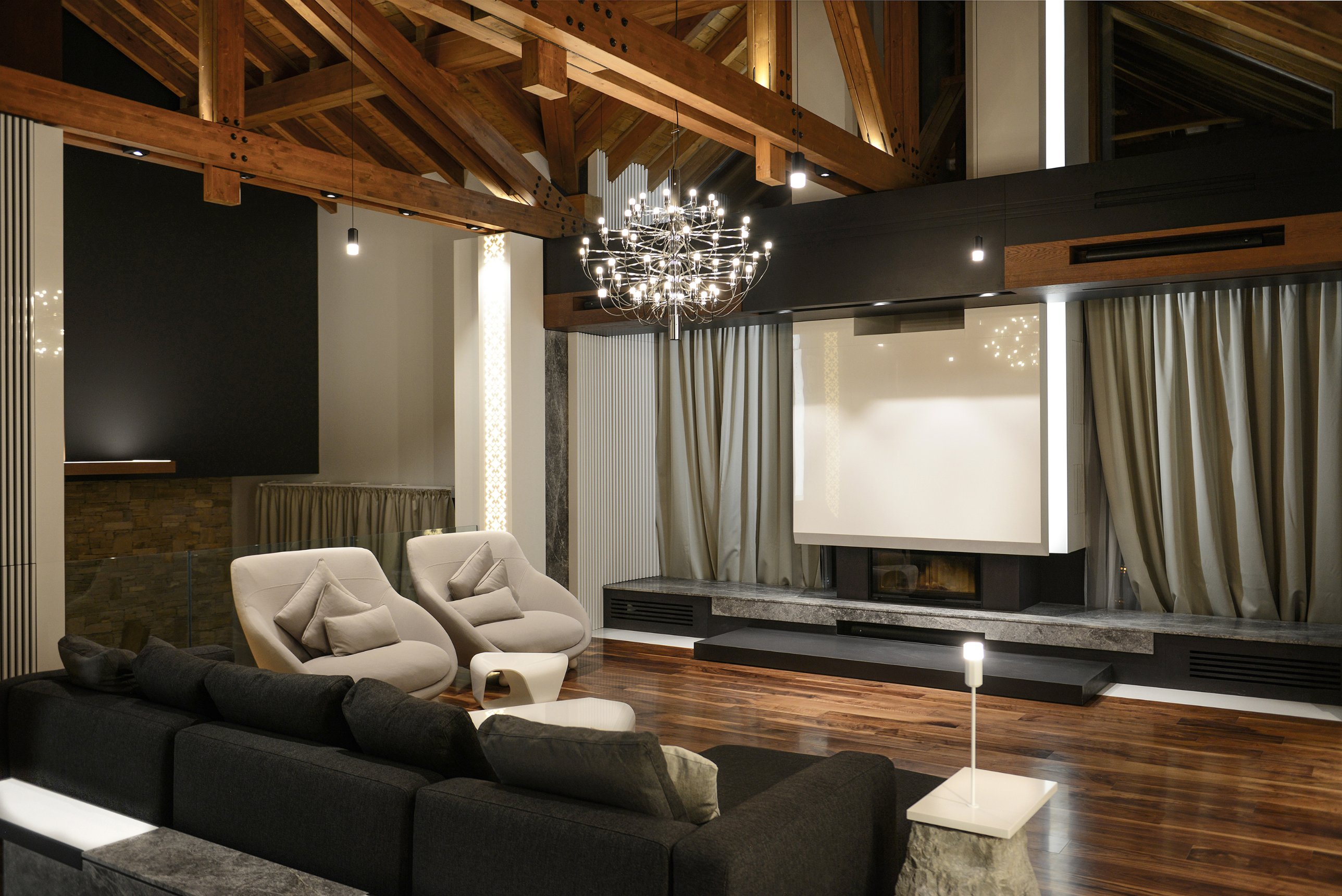 Living room beneath exposed timber trusses, with a chrome chandelier and a stone-based fireplace wall