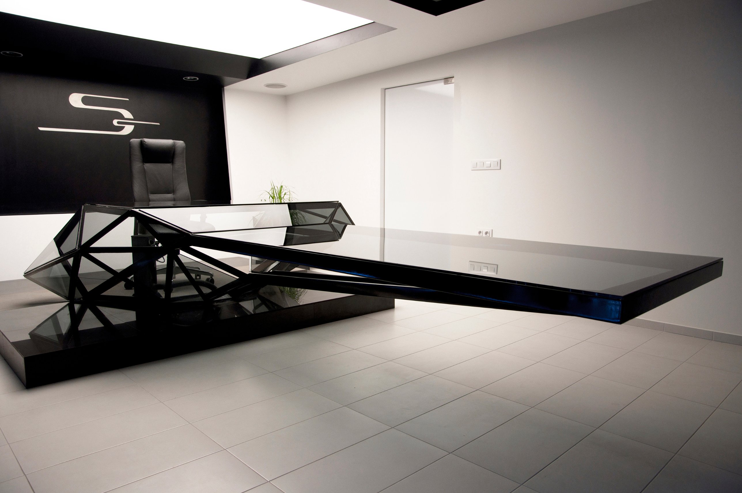 The long glass conference surface cantilevering from the faceted desk across the pale floor