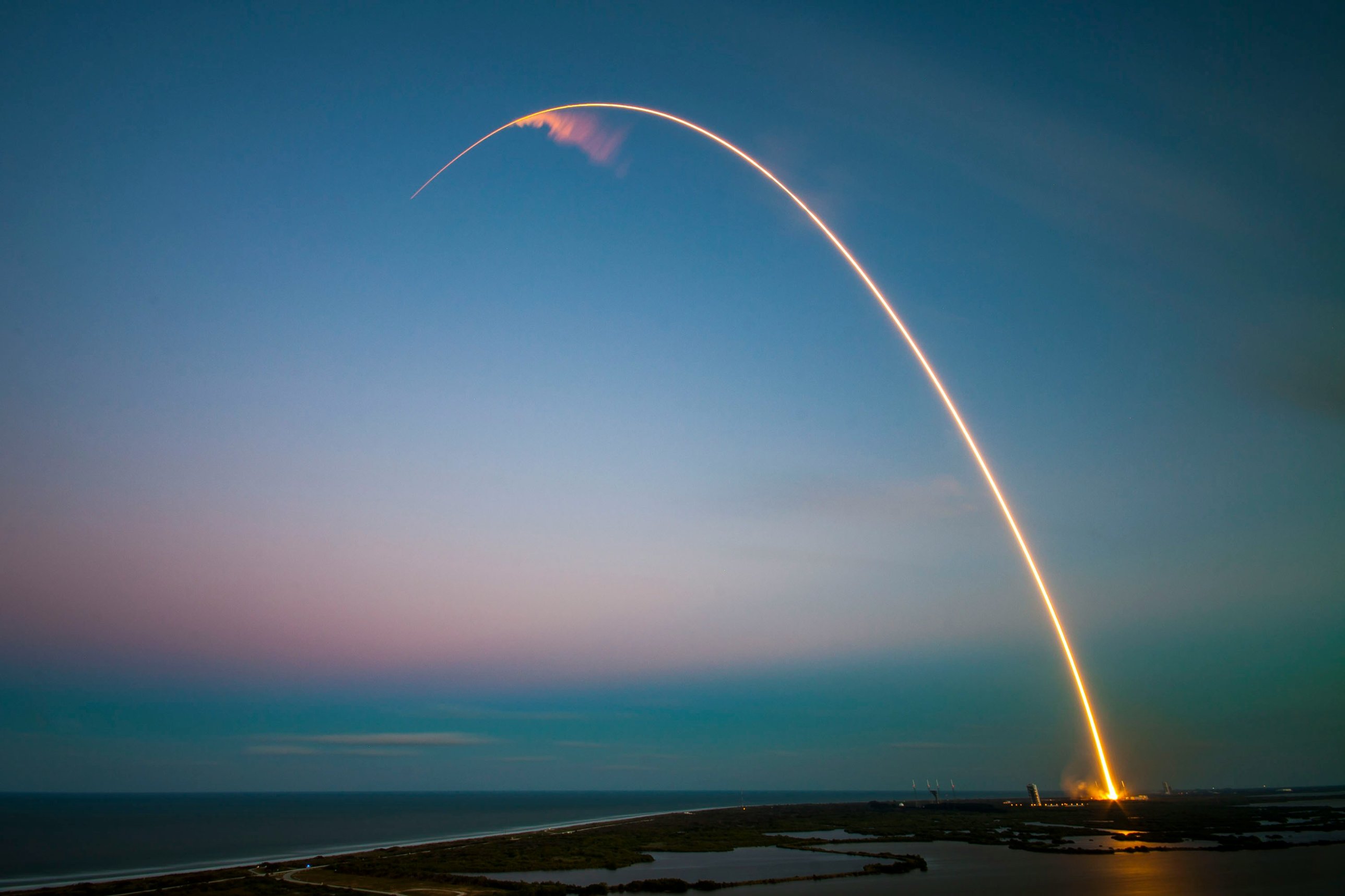 A rocket launch tracing a glowing arc over the coast at dusk